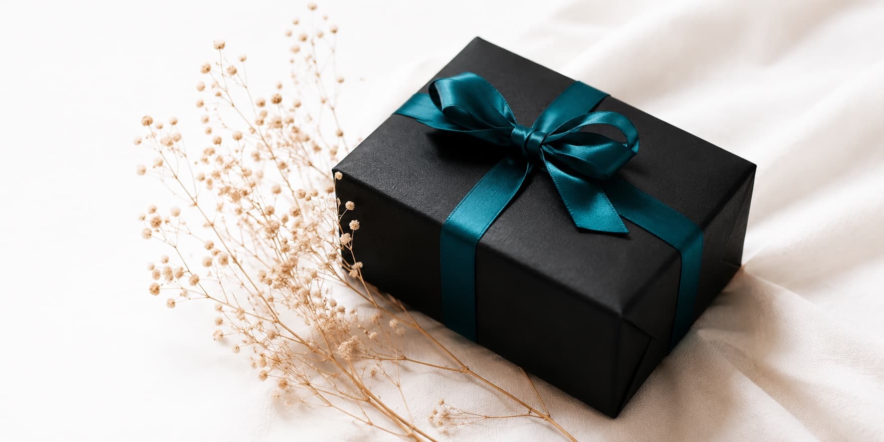Un paquet cadeau noir noué d'un ruban teal, posé sur un tissu clair près de fleurs séchées.